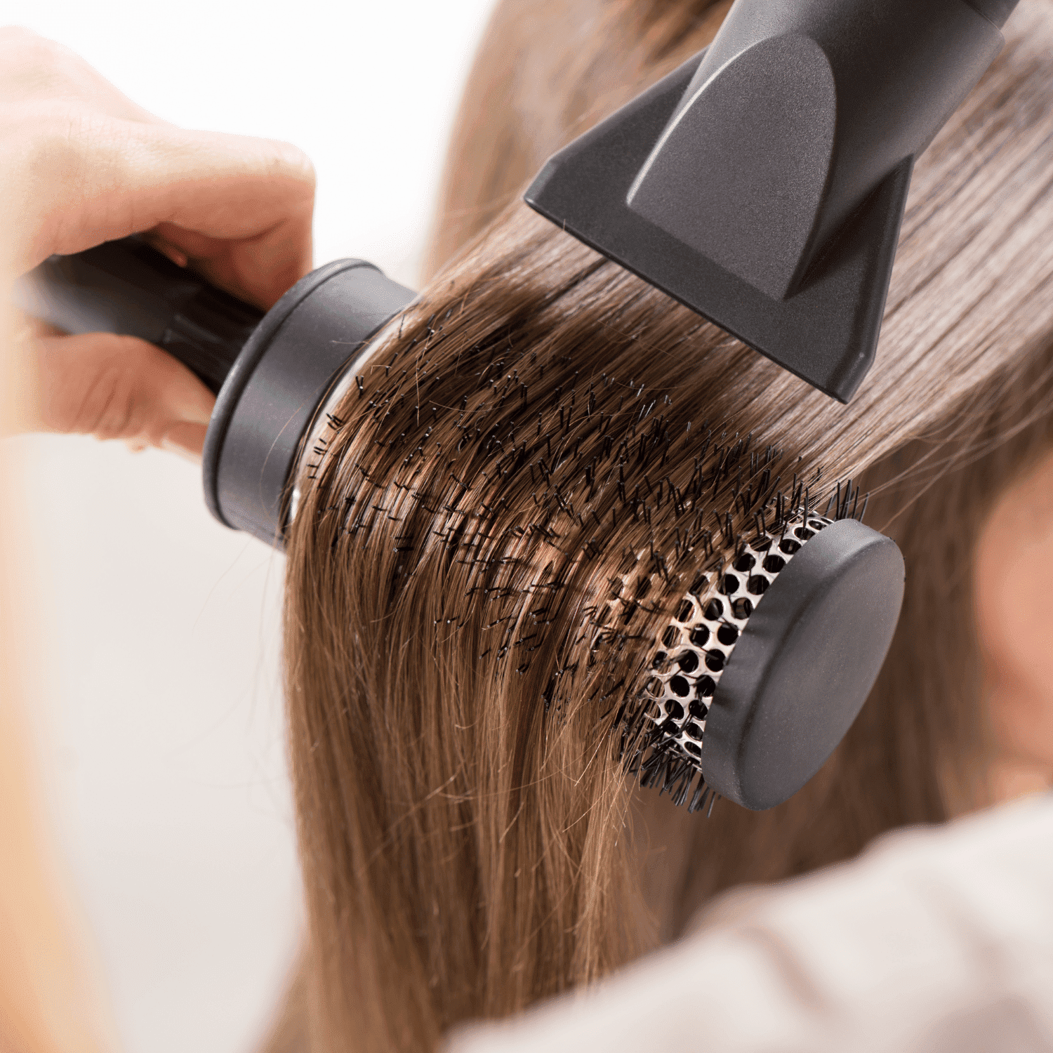 Hands using round brush and hairdryer to style long brown hair.