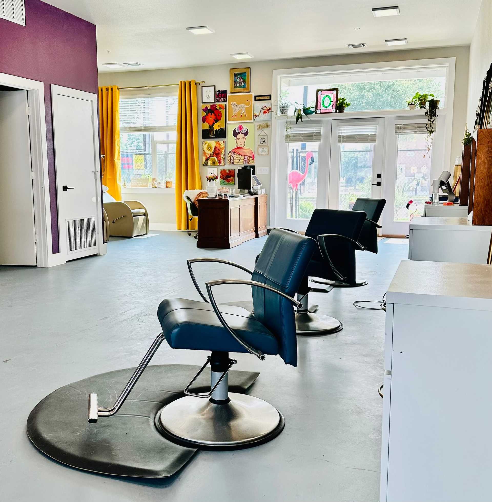 Bright hair salon with styling chairs, colorful wall art, and large windows with yellow curtains.