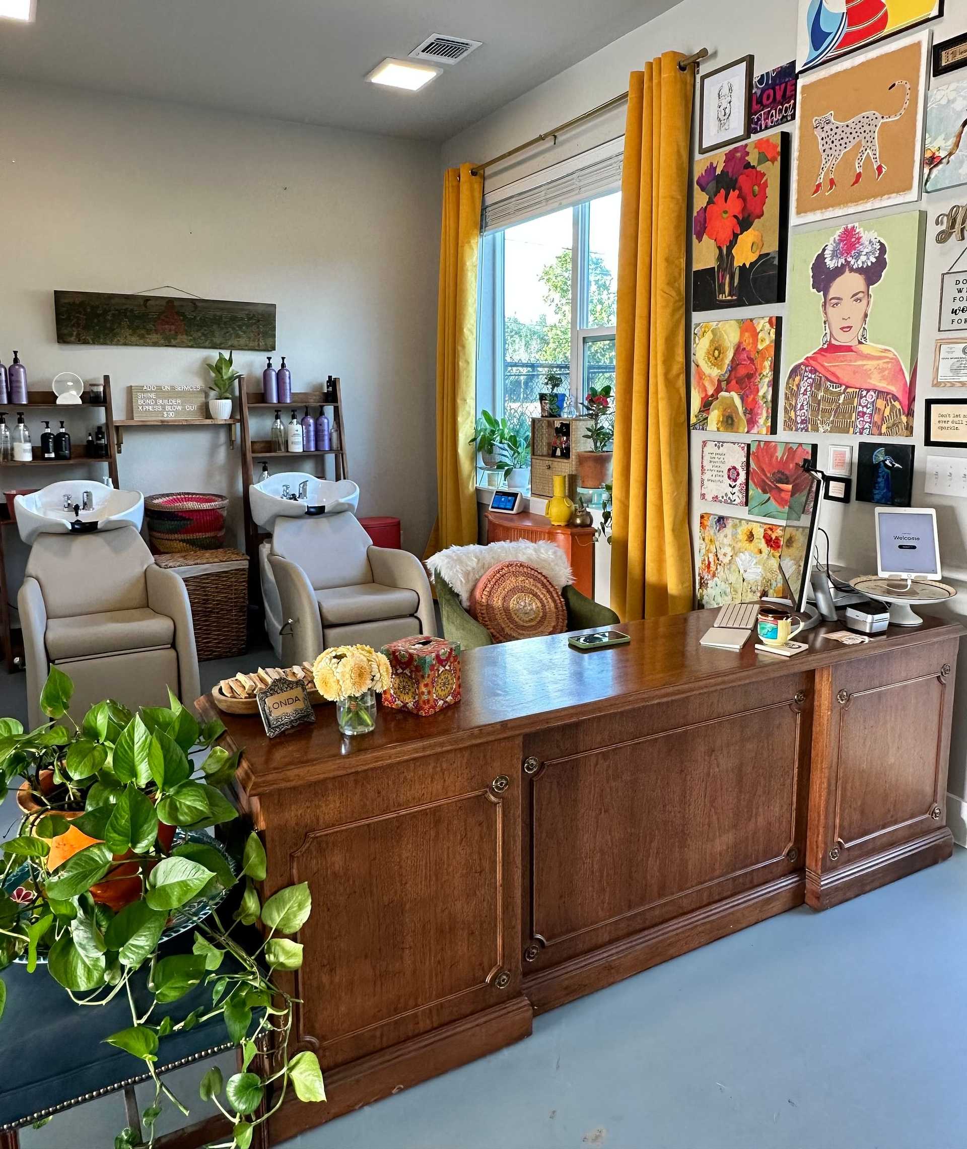 Hair salon with vibrant wall art, cozy chairs, plants, and a wooden desk with floral decorations.